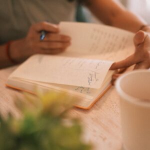 Close-up of a person writing in a notebook with a coffee cup nearby, warm setting.